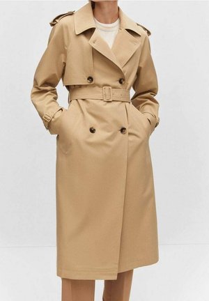 Beige dobbelt-breasted trenchcoat med bælte, knappedetaljer og klap på brystet båret over en hvid top, hænderne i lommerne.