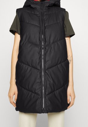 Czarna pikowana bezrękawnik z kapturem, noszona na oliwkowej koszuli z krótkim rękawem, zapinana na zamek, pokazująca tatuaż kreskówkowego króliczka na lewej ręce.