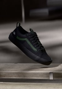 Black sneakers with green accents, lace-up design, rubber sole, and textured fabric. Features a logo on the side and a pull tab on the heel.
