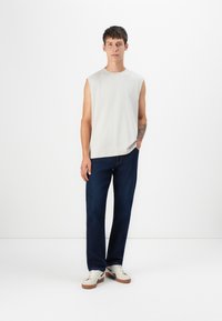 Camisa sin mangas gris, jeans rectos de la marca azul oscuro, y zapatillas color crema con suelas marrones, llevadas por un modelo masculino de pie.