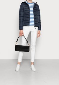 Giacca imbottita blu con cappuccio, camicia a righe blu e bianche, jeans bianchi, borsa nera e sneaker bianche. Texture morbide e design aderente.