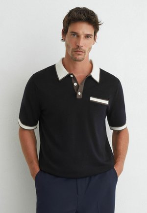 Man wearing a black short-sleeve polo shirt with white collar and pocket trim, standing against a plain white wall with hands in pockets.
