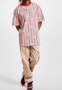 Camiseta de gran tamaño en blanco con rayas verticales rojas y negras. Presenta un diseño de cuello redondo y detalles con texto; combinada con pantalones cargo beige y zapatillas deportivas.