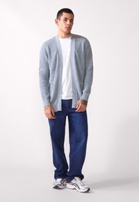 Lys grå strikket cardigan med åpen front, sammen med en hvit t-skjorte og blå jeans, båret med sølvfargede joggesko. Tatovering synlig på armen.
