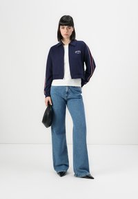 Woman standing wearing a navy zip-up jacket, white shirt, wide-leg blue jeans, black pointed shoes, and holding a black pouch bag.