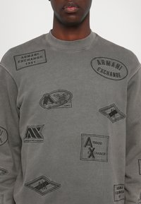 Šedá mikina s různými vyšívanými nášivkami Armani Exchange v černé barvě. Design s kulatým výstřihem, pohodlný střih a texturovaná látka.