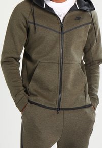 Olivgrön zip-up hoodie med svarta detaljer, med huva, framfickor och Nike-logotyp på bröstet. Mjuk, strukturerad tyg.