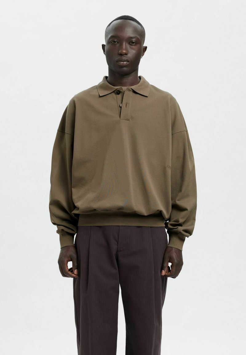 Zaļgana oversize polo džemperis ar apkakli, garām piedurknēm un ribotām aprocēm, kombinēts ar tumši violetiem piegriezuma biksēm.