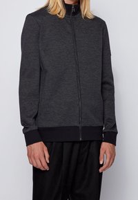 Grå zip-up jacka med hög krage, ribbad mudd och nederkant. Har textilstruktur och kontrasterande svarta detaljer. Inga synliga logotyper.