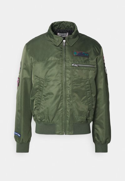 Giacca bomber verde oliva realizzata in nylon liscio con zip frontale, tasche laterali, polsini e orlo a costine, e toppe ricamate sulle maniche.