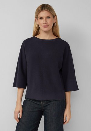 Blondynka ubrana w granatowy prążkowany sweter z łódkowym dekoltem i rękawami o długości trzy czwarte zakończonymi rozszerzeniem, a także ciemnoniebiesje dżinsy, stoi na szarym tle.