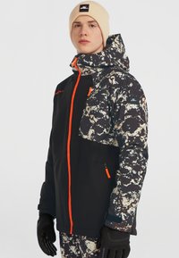 Zwarte en gemusterde sportjacket met een oranje rits, voorzien van een capuchon en zakaccenten. Draagster in zwarte handschoenen en een beige muts.