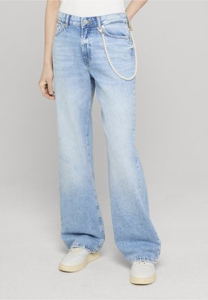 Osoba w jasnoniebieskich, szerokich dżinsach z perełkowym łańcuszkiem i białych tenisówkach, stojąca na tle jednolitego tła.