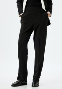 Pantalon noir en tissu lisse et extensible ; doté d'une taille haute, de poches latérales et de lignes épurées ; associé à des mocassins noirs.