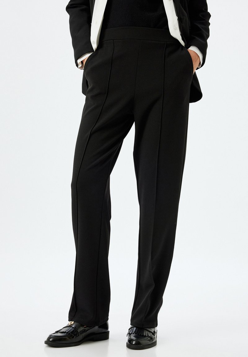 Pantalon noir en tissu lisse et extensible ; doté d'une taille haute, de poches latérales et de lignes épurées ; associé à des mocassins noirs.