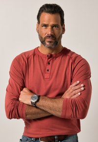Mann mit Bart, der ein rost-rotes Langarmhemd und eine Armbanduhr trägt, steht mit verschränkten Armen vor einem einfarbigen Hintergrund.