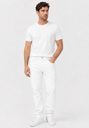 Man draagt een gewoon wit T-shirt, witte spijkerbroek en witte sneakers, staat met één hand in zijn zak tegen een witte achtergrond.