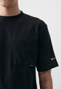 Crna pamučna majica s prednjim džepom, dizajnom s okruglim izrezom i malim logotipom na rukavu. Mekana tekstura i minimalistički stil.