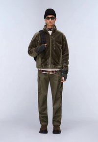 Chaqueta de forro polar verde oliva con cierre frontal, combinada con pantalones cargo verde oliva. Los accesorios incluyen un gorro negro y gafas de sol, llevando una mochila.