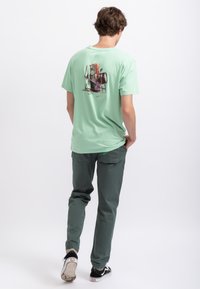 T-shirt en coton vert menthe avec un imprimé graphique coloré au dos. Le porteur l'associe à un pantalon vert foncé et des baskets noires.