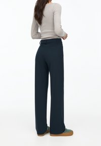Pantalons navy à larges jambes en tissu lisse avec une coupe ajustée, dotés de poches latérales et d'une taille classique. Associés à des baskets vertes.