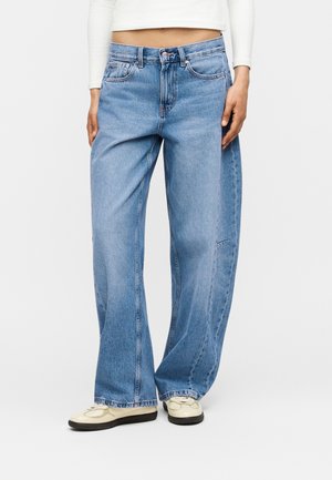 Άτομο που φοράει ανοιχτό μπλε τζιν με φαρδιά μπατζάκια, λευκό cropped μακρυμάνικο τοπ και μπεζ αθλητικά παπούτσια, στέκεται μπροστά σε απλό λευκό φόντο.