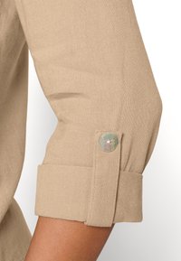 Beżowy rękaw lnianej koszuli z podwiniętym mankietem, ozdobiony metalicznym guzikiem. Tkanina ma teksturowany wygląd i miękkie wykończenie.