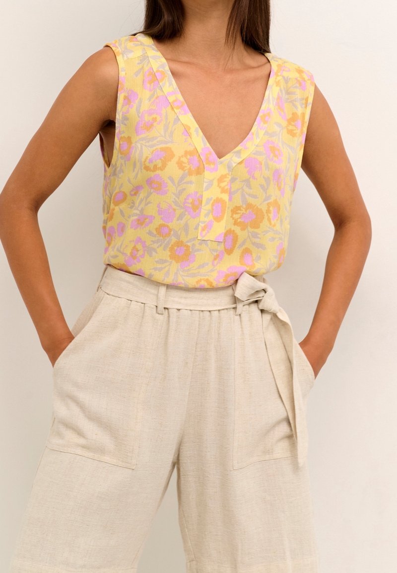 Femme portant un haut sans manches jaune à motif floral et un pantalon beige taille haute avec ceinture nouée, les mains dans les poches sur fond uni.