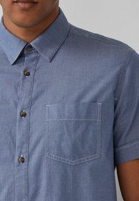 Koszula z krótkim rękawem, zapinana na guziki, w jasnoniebieskim teksturowanym materiale. Posiada kieszonkę na piersi oraz brązowe guziki z kontrastowym wykończeniem szwów.
