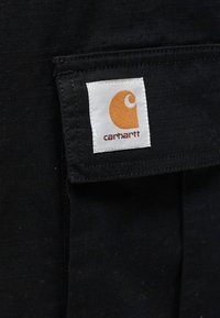 Μαύρο ύφασμα με μοτίβο πλέγματος, με μια λευκή ετικέτα που φέρει πορτοκαλί λογότυπο και την επιγραφή «carhartt», στερεωμένη σε μια τσέπη.