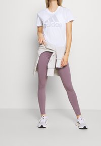 Camiseta blanca de Adidas con logo gris, combinada con leggings morados claros y zapatillas deportivas blancas. Un suéter gris atado a la cintura.