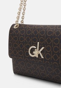 Kabelka Calvin Klein s tmavohnědou látkou se vzorem opakujícího se loga, zlatými kovovými doplňky a řetízkem na rameno. Jednoduchý design s klopou na zavírání.