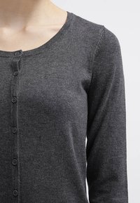 Kvinna som bär en mörkgrå, långärmad, knäppt stickad kofta med rund halsringning mot en enhetlig ljus bakgrund.