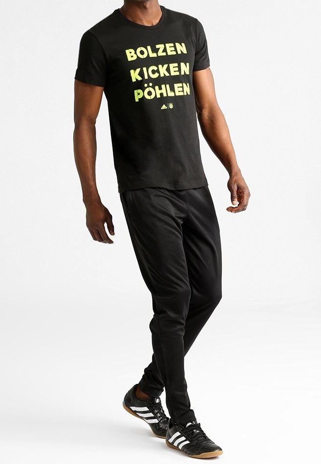 Camiseta negra con el texto amarillo "BOLZEN KICKEN PÖHLEN", pantalones deportivos negros y zapatillas negras con rayas blancas y suela de goma.