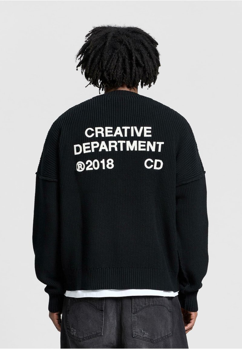 Pull en maille noir au fit oversize, texture côtelée et texte blanc audacieux "CREATIVE DEPARTMENT 2018 CD" dans le dos.