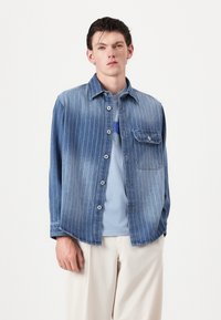 Denim shirt with vertical white stripes, button-up design, single chest pocket, worn over a light blue t-shirt, paired with light trousers.