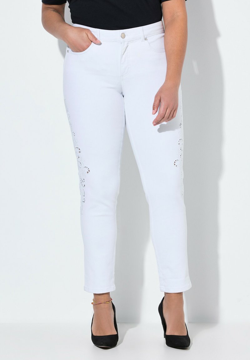Pantaloni in denim bianco con ritagli decorativi lungo i lati, slim fit, lunghezza alla caviglia, abbinati a tacchi alti neri e a una maglietta scura.