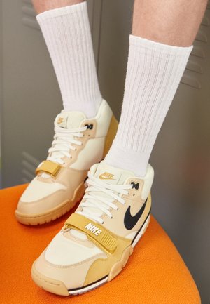 Jambes portant des chaussettes blanches côtelées et des baskets Nike beige avec un logo noir et une bride jaune, reposant sur une surface orange.