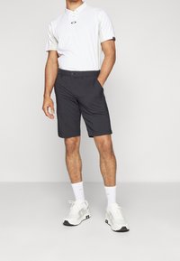 Tenue masculine comprenant un polo blanc avec des accents gris et un short noir. Le short a une coupe classique, un tissu lisse et des poches latérales.