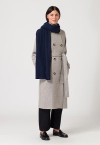 Cappotto di lana grigio con bottoni doppi, cintura coordinata e sciarpa blu navy. Pantaloni neri a gamba larga e scarpe nere completano l'outfit.