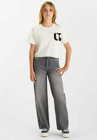 T-shirt blanche courte avec un logo noir sur la poche, associée à un jean en denim gris ample et des baskets bleues. Le modèle se tient devant un fond uni.