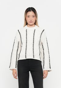 Witte gebreide trui met zwarte gevlochten accenten, kabelbreisel, hoge hals en brede mouwen. Verkorte lengte en textuurstof.