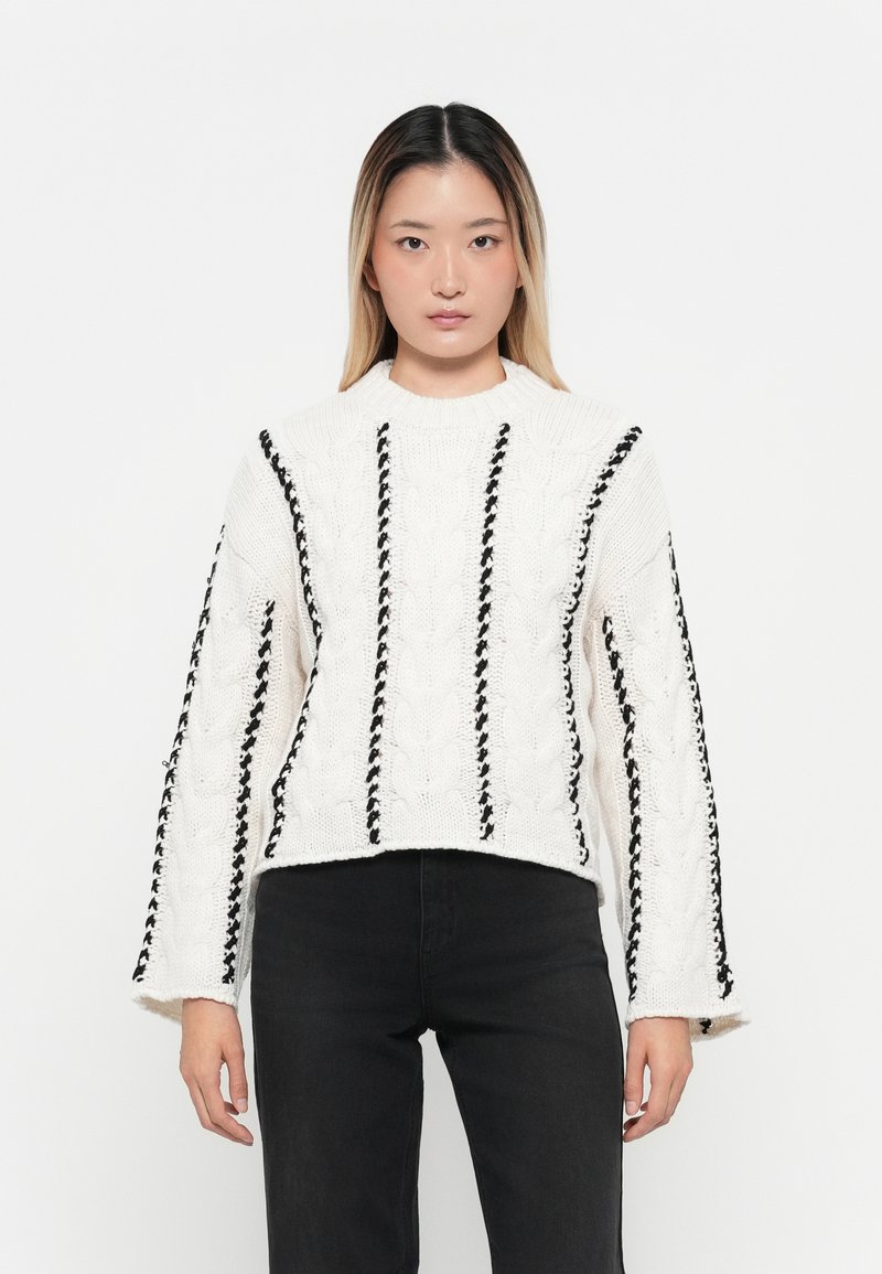 Weißer gestrickter Pullover mit schwarzen geflochtenen Akzenten, Strukturstrickdesign, hoher Kragen und weiten Ärmeln. Kurze Länge und strukturierter Stoff.