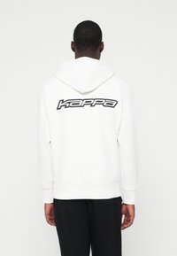 Mann, der einen weißen Hoodie mit einem schwarzen "Kappa"-Logo auf dem Rücken trägt, kombiniert mit schwarzen Hosen, von der Kamera abgewandt.