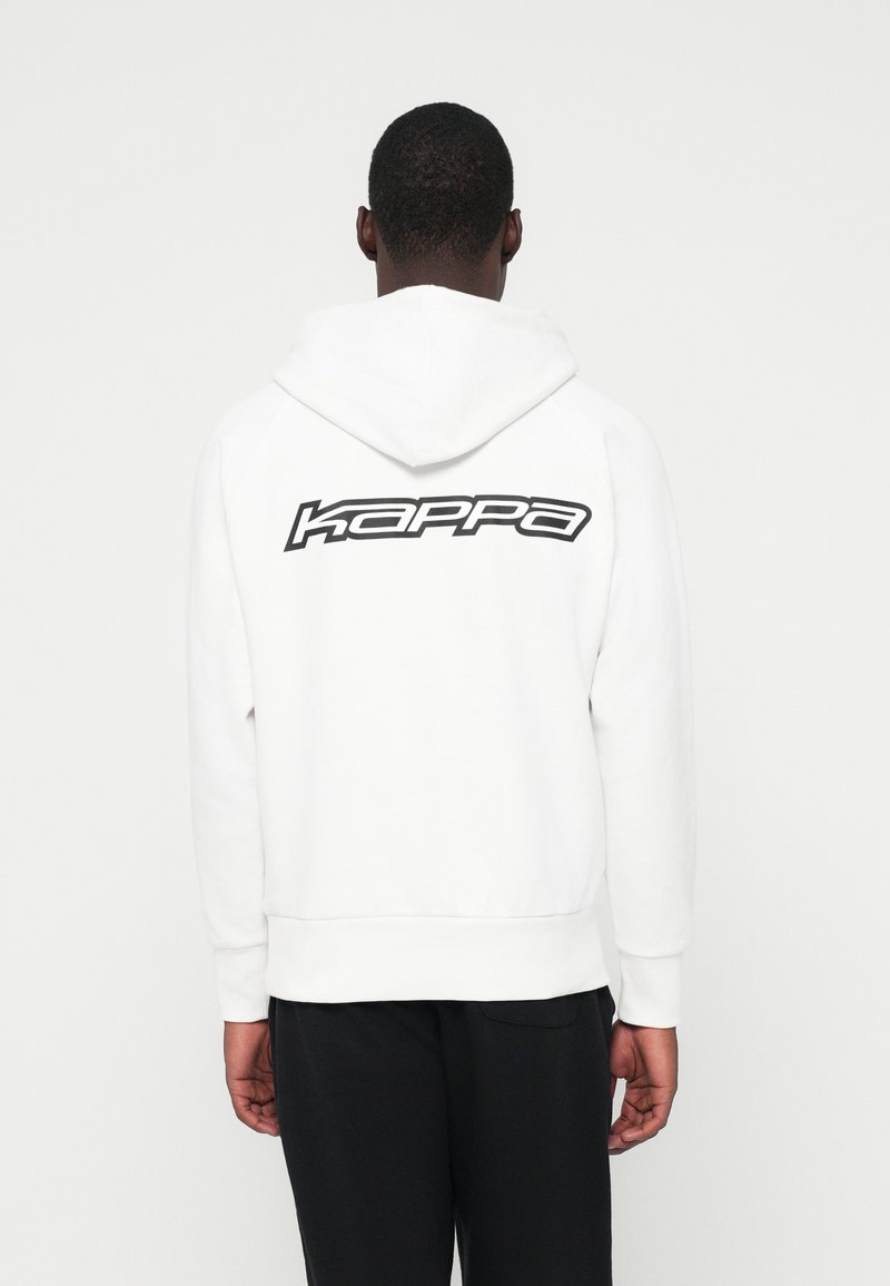 Mann, der einen weißen Hoodie mit einem schwarzen "Kappa"-Logo auf dem Rücken trägt, kombiniert mit schwarzen Hosen, von der Kamera abgewandt.