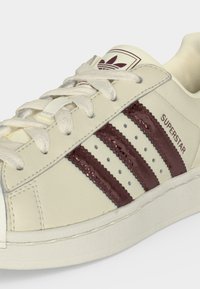 Baskets Adidas Superstar couleur crème avec trois bandes bordeaux, lacets blancs et inscription « SUPERSTAR » sur la tige côté.