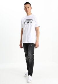 Weißes Grafik-T-Shirt mit schwarzem Vans-Logo, kombiniert mit zerrissenen, dunkelgrauen Jeans und weißen Sneakers, was ein lässiges Outfit ergibt.