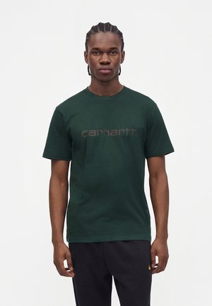 Jeune homme aux cheveux tressés portant un T-shirt Carhartt vert foncé et un pantalon noir, debout devant un fond blanc uni.