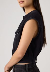 Νεαρή γυναίκα με μακριά σκούρα μαλλιά, που φοράει αμάνικο μαύρο τζιν γιλέκο και μαύρα τζιν, εμφανίζεται από το πλάι σε απλό φόντο.