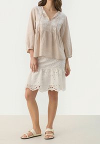 Beigenvärinen pellavapusero, jossa on kukkakirjailu, yhdistetty valkoiseen, reikäkohosomisteiseen pitsihameeseen ja beigenvärisiin liukusandaaleihin. Rentoutunut istuvuus ja levenevät hihat.